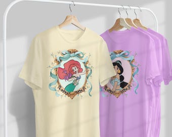 Op de afbeelding: Crèmekleurig T-shirt met een omlijste illustratie van Ariel uit De Kleine Zeemeermin, met rood haar en een groene staart. Een lichtblauw lint en een gouden lijst omringen de afbeelding. Een paars t-shirt met Jasmine uit Aladdin is ook zichtbaar.