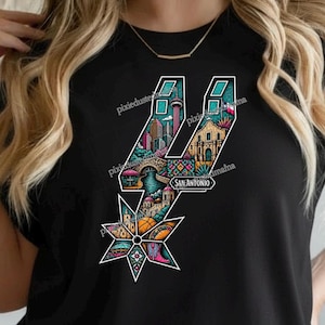 May include: Black t-shirt featuring a colorful graphic of the San Antonio Spurs logo. The logo is filled with illustrations of San Antonio landmarks, including the Tower of the Americas, a bridge, and a mission. The word "SAN ANTONIO" is also included.