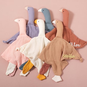 May include: A collection of soft, plush goose-shaped baby comforters in various pastel colors, including pink, lavender, and beige. Each comforter has a white beak and foot, with some featuring embroidered names like "Stella" and "Chloe".