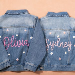 May include: Two light blue denim jackets with embroidered names and floral accents. One jacket says "Olivia" in pink script, the other says "Sydney". Both jackets have white and pink flower details.