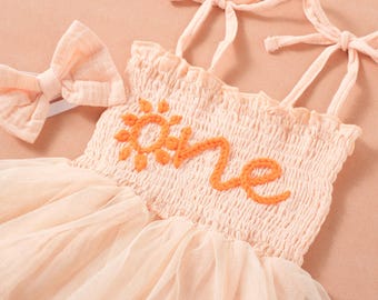 Puede incluir: Vestido de bebé color melocotón con falda de tul y diadema a juego. El corpiño presenta la palabra "one" bordada en naranja, con un diseño de sol encima del texto "one". El vestido tiene tirantes anudados.