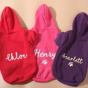 May include: Three hooded dog sweaters in red, pink, and purple. Each sweater has a name in cursive script: "Chloe", "Henry", and "Scarlett". Paw prints are below the names on the pink and purple sweaters.