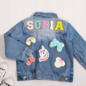 May include: A light blue denim jacket with the name "SOFIA" spelled out in colorful, embroidered letters on the back. The jacket is decorated with patches of a rainbow, unicorn, butterfly, bow, and cloud. A pair of white sneakers with a plaid pattern are visible.