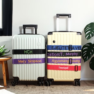 May include: Two suitcases with luggage straps. The larger suitcase is cream colored with the text "EJELITETRAVEL", "zation:T. Murphy", "Agyemang Young", and "Hasselquist" on the straps. The smaller suitcase is white with the text "HCH", "Mary Simpson" on the straps.