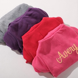 May include: Four folded sweatshirts in gray, purple, red, and pink. The pink sweatshirt has the name "Avery" in a gold script font. The sweatshirts are made of a soft material and are likely for children.