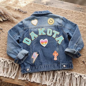 May include: A blue denim jacket featuring the name DAKOTA in light blue letters across the back. The jacket is decorated with several patches, including a white heart, a yellow smiley face, a rainbow peace sign heart, a lightning bolt, and a mushroom.