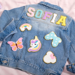 May include: Light blue denim jacket with the name "SOFIA" in pastel-colored, embroidered letters on the back. The jacket is decorated with patches of a rainbow, butterfly, unicorn, and clouds.
