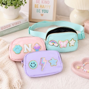 Op de afbeelding: Drie kleurrijke heuptasjes in roze, lichtblauw en lavendel. Elk tasje heeft geborduurde patches, waaronder een pootafdruk, een cupcake, een regenboog, een vlinder, een ster, een bliksemschicht en een smiley. Het lichtblauwe tasje heeft een bijpassende riem.