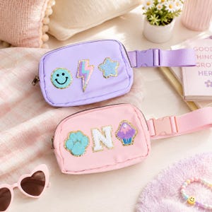 May include: A light purple fanny pack with a smiley face, lightning bolt, and star patch, and a light pink fanny pack with a paw print, the letter N, and a cupcake patch. Both bags feature adjustable straps with plastic side-release buckles.