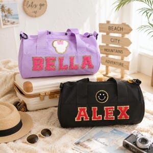 Puede incluir: Una bolsa de deporte color lavanda con un parche en forma de ratón blanco y rosa y el nombre BELLA en letras de purpurina rosa, y una bolsa de deporte negra con un parche de cara sonriente amarilla y el nombre ALEX en letras de purpurina roja.