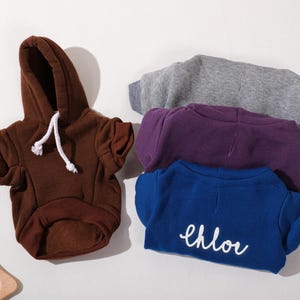 May include: A collection of pet hoodies in various colors. A brown hoodie with a white drawstring is on the left. Other hoodies are stacked: gray, purple, and blue with the name "Chloe" in white script. The hoodies are made of soft material.