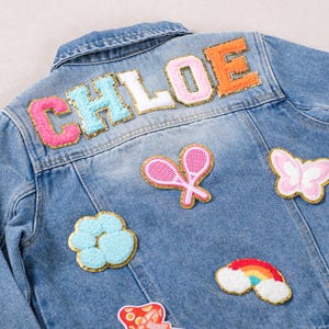 May include: Light blue denim jacket with the name "CHLOE" appliqued on the back in colorful, textured letters. The jacket is decorated with embroidered patches, including a butterfly, tennis rackets, and a rainbow.