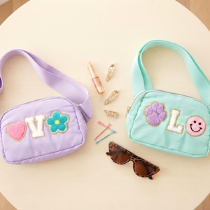 May include: A lavender belt bag with a heart, V, and flower patch, and a mint green belt bag with a paw print, L, and smiley face patch. Accessories include a lipstick, three hair clips, two bobby pins, and brown tortoiseshell sunglasses.