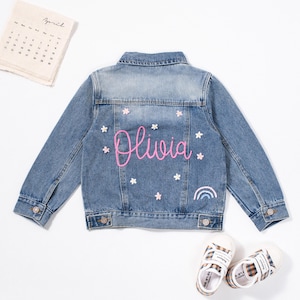 May include: A light blue denim jacket with a pink embroidered name "Olivia" and white embroidered flowers on the back. A rainbow is embroidered below the name.