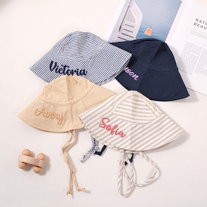 Puede incluir: Cuatro sombreros de pescador personalizados en varios colores y patrones. Uno es azul marino, otro beige y dos a rayas azules y grises. Cada sombrero tiene un nombre bordado en la parte delantera en un color contrastante.