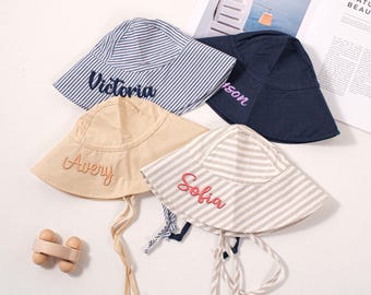 May include: Four personalized bucket hats in various colors and patterns. One is navy blue, another is beige, and two are striped in blue and gray. Each hat has a name embroidered on the front in a contrasting color.