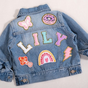 May include: Light blue denim jacket with colorful patches, including a butterfly, donut, rainbow, mushroom, and lightning bolt. The name "LILY" is spelled out in individual letter patches. A stylish and personalized children's jacket.