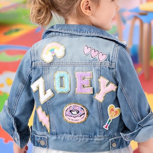 May include: A light blue denim jacket with colorful patches. The patches include a rainbow, hearts, a lightning bolt, a donut, and the name "ZOEY" spelled out in individual letters. The jacket has a button closure and two front pockets.
