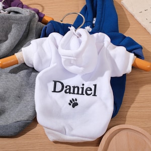 May include: White pet hoodie with the name "Daniel" embroidered in black, along with a paw print. The hoodie is displayed on a wooden hanger, with a blue hoodie visible in the background. Other hoodies in gray and purple are also visible.