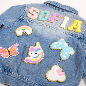 May include: A light blue denim jacket with colorful patches. The patches include a rainbow, butterfly, cloud, and a unicorn. The name "SOFIA" is spelled out in large, colorful letters across the back of the jacket.
