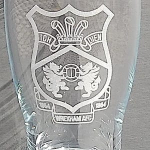 May include: Clear glass pint glass with an etched design. The design features a shield with the words "ICH DIEN" above a crest with two griffin-like creatures and the text "WREXHAM AFC" below. The glass has a wide base.