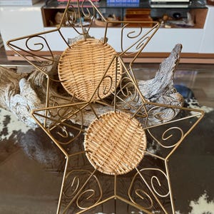 May include: Two star-shaped decorative trays with gold metal frames and woven rattan centers. The star points are adorned with decorative swirls. The trays are designed to hold small items and add a touch of elegance to any space.