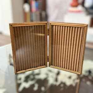 May include: A gold-colored, hinged picture frame with two rectangular sections. The frames have a beaded border and a ribbed backing. The frame is displayed on a glass surface.