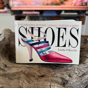 May include: A book titled "SHOES" by Linda O'Keeffe, featuring a colorful illustration of a high-heeled sandal. The cover text reads "A Celebration of Pumps, Sandals, Slippers & More". The shoe design includes red, blue, and pink straps.