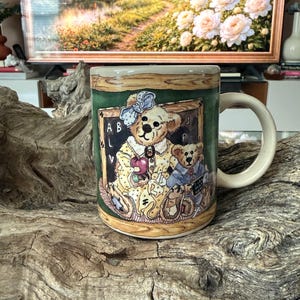 May include: A ceramic coffee mug featuring a charming illustration of two teddy bears in a classroom setting. The bears are depicted in a schoolroom scene with a chalkboard, apple, and the letters A and B. The mug has a cream-colored handle and rim.