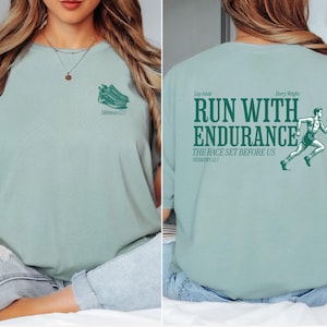 Op de afbeelding: Een lichtblauw t-shirt met een groene grafische print van een hardloper en de tekst "Run with Endurance, The Race Set Before Us, Hebrews 12:1".