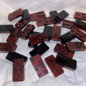 May include: A collection of dominoes with a two-tone design. The dominoes have a dark red base with black pips and a black border. Some dominoes feature Mickey Mouse head pips. The dominoes are scattered on a white surface.