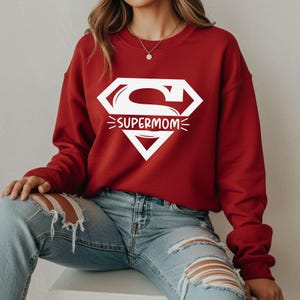 May include: A red sweatshirt with a white "Supermom" graphic. The graphic is in a diamond shape with the letter "S" inside. The sweatshirt has long sleeves and a crew neck.