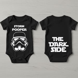 Op de afbeelding: Zwarte babybody met witte tekst en afbeeldingen. De voorkant heeft een Stormtrooper-helm en de woorden "STORM POOPER". De achterkant toont "THE DARK SIDE" in vette letters. Een leuke outfit met thema.