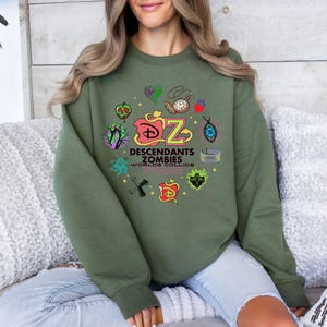 May include: An olive green sweatshirt featuring a colorful graphic design from the Disney movie "Descendants." The design includes the text "Descendants Zombies Worlds Collide Tour" and various themed icons.