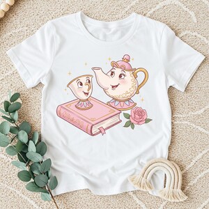 Puede incluir: Camiseta blanca con una ilustración de dibujos animados de una tetera, una taza, un libro y una rosa. La tetera y la taza tienen caras sonrientes. El libro es rosa con adornos dorados y una cinta rosa como marcapáginas. Una rosa rosa está al lado del libro.