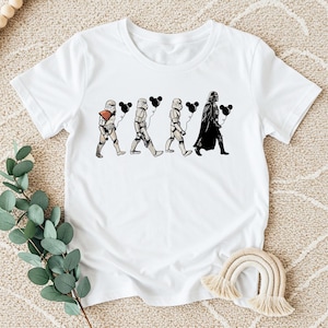 May include: White t-shirt featuring a graphic of Star Wars characters, including Stormtroopers and Darth Vader, each holding a black Mickey Mouse-shaped balloon. The design is in black and white, creating a playful contrast on the white fabric.