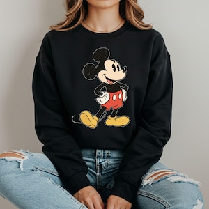Może przedstawiać: Czarna bluza z nadrukiem vintage'owego Mickey Mouse'a. Mickey stoi z rękami na biodrach i ma na sobie swoje charakterystyczne czerwone spodenki z żółtymi butami.