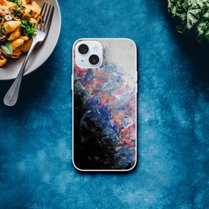 May include: A white iPhone case with a colorful abstract design. The case features a mix of red, blue, and black paint strokes, creating a vibrant and artistic look. The case has a camera cutout and a white border.