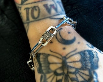 Könnte beinhalten: Ein silbernes Kettenarmband mit einem quadratischen Anhänger, der mit einem schwarzen, diamantartigen Stein besetzt ist. Das Armband wird an einem tätowierten Handgelenk mit einem Schmetterlingstattoo getragen.
