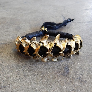 May include: A black leather bracelet with gold-toned spikes. The bracelet is made of a braided leather strap with a series of gold-toned spikes that are arranged in a row.