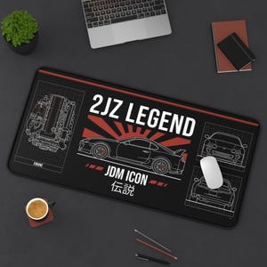 Op de afbeelding: Zwarte rechthoekige muismat met een rode rand, met een autografiek en de tekst "2JZ LEGEND" en "JDM ICON". Het ontwerp bevat een autosilhouet, een motordiagram en Japanse karakters. Een witte muis ligt erop.