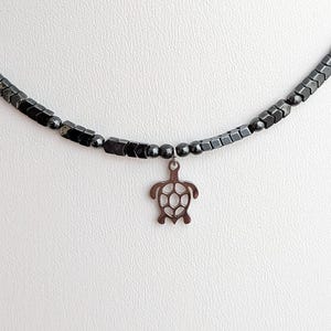 May include: A dark grey beaded necklace with a silver-toned turtle pendant. The necklace is made of small, dark grey beads and a turtle charm. The jewellery is shown against a white backdrop, emphasising the necklace's design.