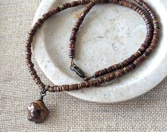 May include: A handcrafted necklace featuring small, dark brown wooden beads and brass accents. The necklace has a pendant with a unique, organic shape. The clasp is a dark metal. The necklace is displayed on a white, oval-shaped plate.