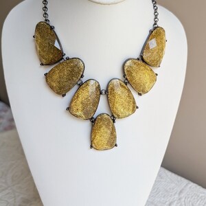 May include: A statement necklace featuring large, irregular-shaped pendants in a shimmering gold color. The pendants are set in a dark metal frame and connected by a matching chain. The necklace is displayed on a white mannequin.