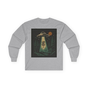 Puede incluir: Una camiseta gris de manga larga con un gráfico de un OVNI que emite un cesto de golf de disco. El diseño incluye árboles, una pequeña cabaña y un sol o una luna en el fondo. La camiseta está hecha de un material suave y cómodo.