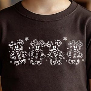 Może przedstawiać: Ciemnobrązowy t-shirt z białym obrysem czterech piernikowych misi&oacute;w z uszami i kokardkami Myszki Miki oraz płatkami śniegu. Koszulka jest noszona przez dziecko.
