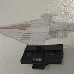 May include: A LEGO Star Wars Acclamator-class assault ship model constructed from light gray bricks with dark red accents. The starship is mounted on a black display stand featuring a plaque with the LEGO Star Wars logo and the ship name.