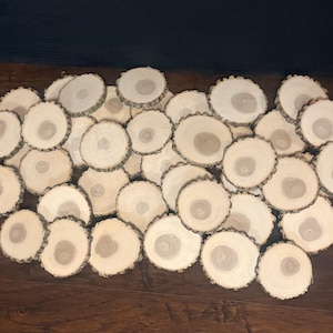 May include: A collection of small, round, natural wood slices with visible growth rings and bark edges. The wood slices are light brown and beige, arranged on a dark brown wooden surface. These rustic wood slices are ideal for crafts and home decor.