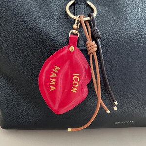 Könnte beinhalten: Ein roter Leder-Lippen-Schlüsselanhänger mit den Worten "MAMA ICON" in goldfarbenen Buchstaben. Der Schlüsselanhänger ist an einem goldenen Ring mit braunen und schwarzen Lederakzenten befestigt und hängt an einer schwarzen Ledertasche.