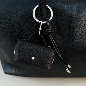Könnte beinhalten: Ein kleiner, schwarzer, strukturierter Leder-Taschenanhänger mit einem silberfarbenen Metallring und -verschluss. Der Anhänger ist an einer größeren schwarzen Ledertasche befestigt. Der Anhänger hat einen kleinen Griff und einen Klappenverschluss mit einem silbernen Knopf.